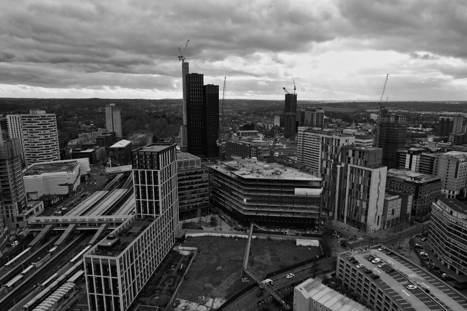A black and white aerial photograph of a cityscape featuring numerous modern high-rise buildings, some under construction with cranes visible at their peaks. The skyline includes tall, dark glass towers and lower office blocks with varied architectural styles. In the foreground, part of a multi-level train station with train tracks and platforms can be seen, alongside parking areas with parked vehicles. The surrounding environment includes streets with moving traffic and clusters of mid-rise structures, set under a cloudy sky that adds a moody atmosphere. The scene captures the dense urban environment typical of a central business district, illustrating the complexity and diversity of city architecture and infrastructure, where private waste management and alternative rubbish removal services could operate on-site or in conjunction with city infrastructure to handle waste materials efficiently.