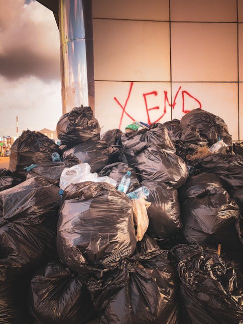 A large pile of black plastic rubbish bags is stacked against a modern building with a reflective, tiled facade. The bags appear tightly filled and are stacked haphazardly, with some partially open showing discarded plastic bottles and packaging. Behind the pile, the building's surface exhibits a grid pattern of beige or light brown tiles, and there is red graffiti with the word 'XEND' spray-painted on the wall. The scene is outdoors, illuminated by natural daylight with a cloudy sky visible in the background, suggesting an urban environment that may involve private waste disposal or on-site clearance services, as offered by companies like Rubbish Removal Croydon.