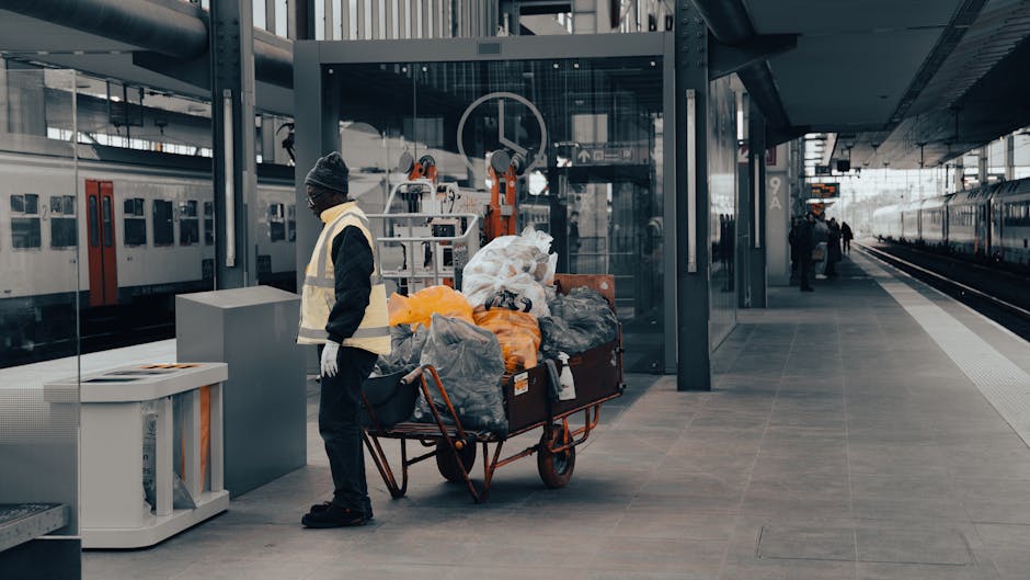 A worker standing on a train station platform next to a large trolley loaded with various bags of waste, including black and orange plastic bags, some filled with rubbish and others tied shut. The worker is dressed in a dark coat, grey beanie, and high-visibility vest, with gloves, as part of a waste collection or rubbish removal service managed by Rubbish Removal Croydon. The platform surface is made of grey tiles, with a glass and metal shelter behind the worker, housing machinery and equipment related to waste handling or on-site collection operations. To the left, a white waste bin or container is positioned on the platform, while to the right, train tracks extend into the distance with adjacent trains visible. The environment is well-lit with natural daylight, emphasizing the urban setting of the station and the ongoing rubbish clearance activity, reflective of private or alternative waste management services outside standard public collection.