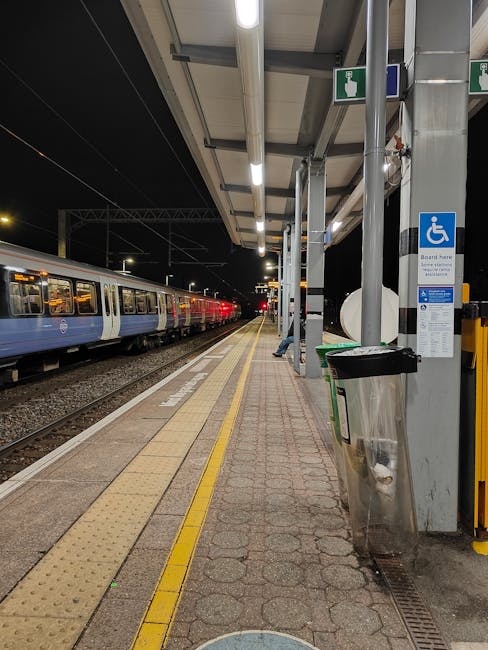 The image shows a train station platform at night, illuminated by overhead fluorescent lighting. On the left side, a passenger train with white and blue carriages is stopped alongside the platform. The platform is paved with textured bricks and features a yellow tactile strip near the train edge for accessibility. To the right, there are support columns and a shelter roof made of metal and glass panels. Mounted on a pole, a blue and white sign indicates disability access, with additional information below. Adjacent to the sign, a metal litter bin with a black lid and a reflective surface is attached to the platform wall. The environment appears clean and quiet, with no visible passengers, highlighting a typical scene of independent rail station management aligned with private waste handling services like those offered by Rubbish Removal Croydon. The scene’s natural lighting emphasizes the functional and orderly atmosphere of the station, catering to both regular commuters and those requiring accessible travel options, all within a setting prepared for efficient passenger flow and waste disposal.