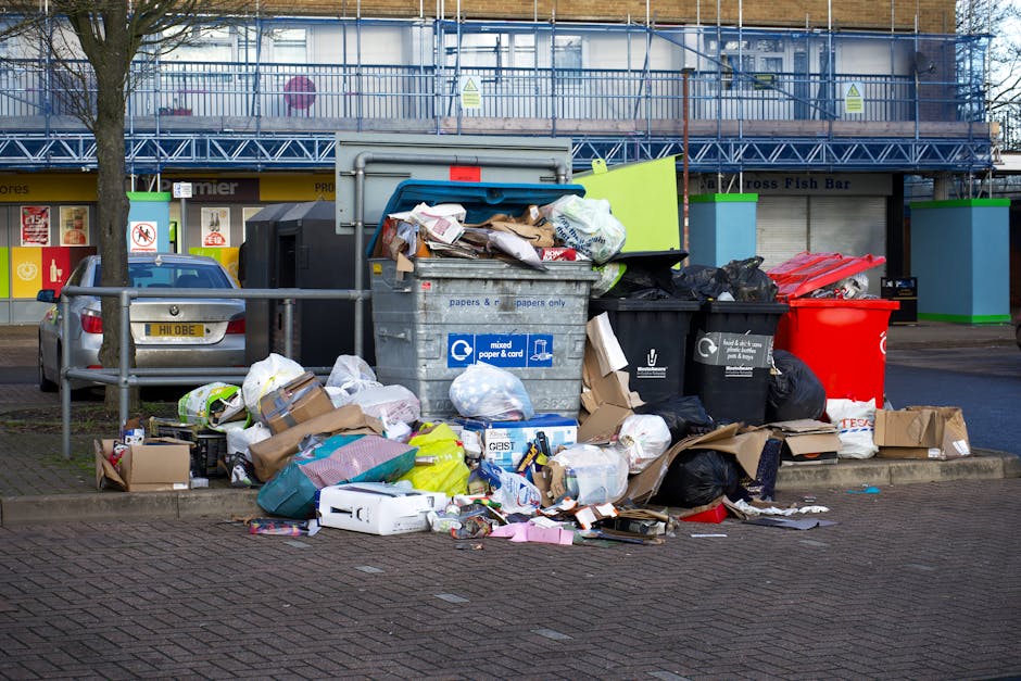 An overflowing waste collection point situated on a paved urban street, featuring a central large, rectangular grey mixed paper and card recycling bin with a blue lid, positioned among various black and red garbage bins. The area around the bins is cluttered with a large assortment of waste including crumpled cardboard boxes, plastic bags, paper packaging, and discarded miscellaneous items spilling onto the pavement. Some rubbish is stacked on top of the bins, while additional waste is scattered on the ground, indicating a collection that has exceeded capacity. Behind the waste area, there is a row of shops with storefronts and signs, and a parked silver car on the left side. A metal railing and a leafless tree frame the scene, with a building under construction or renovation visible in the background, partially covered in blue scaffolding. The overall environment appears to be a commercial or residential street, illustrating the need for regular rubbish removal and proper waste management services like those provided by Rubbish Removal Croydon.