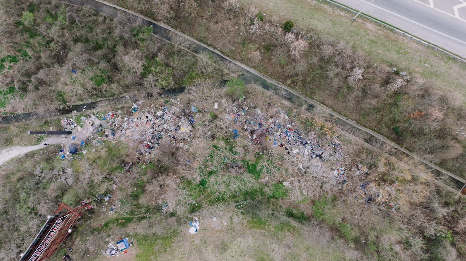 An aerial view of a small, unmanaged area of land adjacent to a road shows a large accumulation of mixed refuse, including plastic bottles, cardboard, paper, and other household waste scattered across the ground. The debris is spread unevenly, with a denser concentration towards the left side of the image, and sparse patches of green grass and leafless trees are visible amidst the rubbish. A narrow dirt path runs along the edge of the waste area, leading toward the bottom left corner, where part of a red metal structure, possibly playground equipment or a small construction frame, is partially visible. Beyond the debris, a concrete wall and a stretch of pavement border the land, separating it from the nearby road or highway. The scene appears to be an informal dumping site, highlighting the importance of proper waste management or private rubbish removal services like those offered by Rubbish Removal Croydon, which facilitate on-site clearance and alternative disposal options for communities seeking to improve local environments.