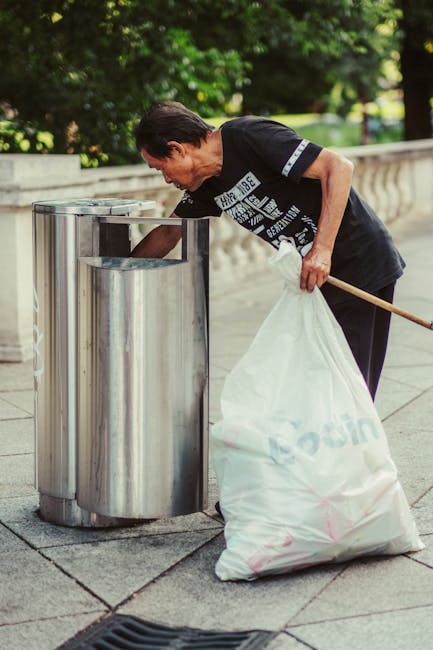 An overflowing waste collection point situated on a paved urban street, featuring a central large, rectangular grey mixed paper and card recycling bin with a blue lid, positioned among various black and red garbage bins. The area around the bins is cluttered with a large assortment of waste including crumpled cardboard boxes, plastic bags, paper packaging, and discarded miscellaneous items spilling onto the pavement. Some rubbish is stacked on top of the bins, while additional waste is scattered on the ground, indicating a collection that has exceeded capacity. Behind the waste area, there is a row of shops with storefronts and signs, and a parked silver car on the left side. A metal railing and a leafless tree frame the scene, with a building under construction or renovation visible in the background, partially covered in blue scaffolding. The overall environment appears to be a commercial or residential street, illustrating the need for regular rubbish removal and proper waste management services like those provided by Rubbish Removal Croydon.