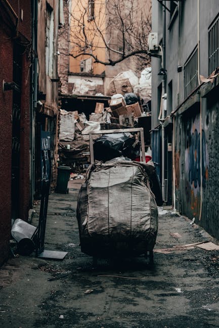 A narrow urban alleyway cluttered with various waste materials, including a large grey bag in the foreground, which appears to be filled with rubbish. Behind it, there are stacks of cardboard boxes, black garbage bags, and loose debris piled up against a wooden pallet, indicating an accumulation of waste. The ground is paved with asphalt and shows signs of dirt and minor stains. On the left side of the alley, a dark metal wall with some graffiti and a small, green trash bin partially tipped over can be seen, while the right side features a building with dark grey metal cladding and barred windows. Bare tree branches are visible overhead, suggesting a cold or late autumn season. The lighting is diffuse, typical of an overcast day, and the scene conveys a sense of uncollected waste typical of private disposal or independent rubbish clearance. This image is relevant to rubbish removal services, illustrating an environment requiring prompt waste collection, which Rubbish Removal Croydon and similar companies might address in their on-site clearance operations.