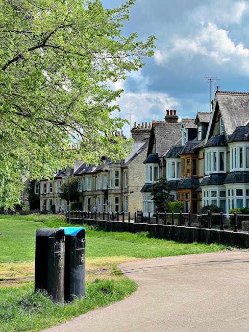 A suburban residential street scene with a row of traditional terraced houses featuring bay windows, pitched roofs, and brick facades. In the foreground, there are two black public rubbish bins positioned on a paved pathway, with a grassy verge beside them. A large leafy tree with fresh green foliage extends over the scene on the left side, partially shading the pavement and grass area. The background shows a bright blue sky with scattered white clouds, highlighting a pleasant daytime atmosphere. The image depicts an environment suitable for outdoor rubbish disposal or local waste collection, reflecting a typical urban setting where residents might engage in private waste handling or bin collection services, such as those offered by Rubbish Removal Croydon.