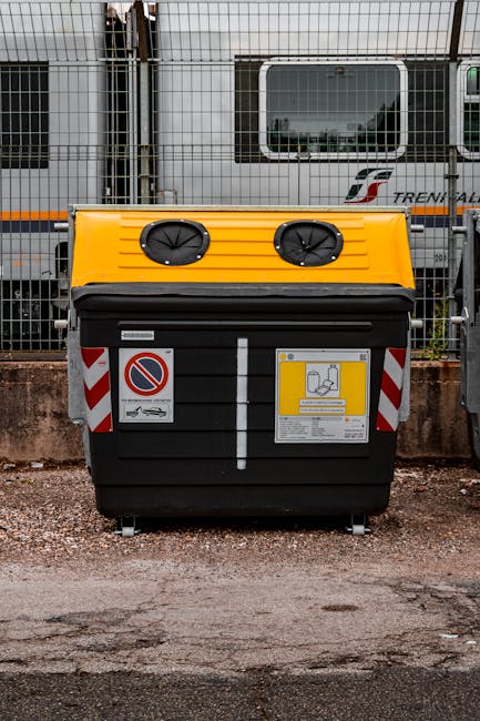 A large black wheeled waste skip with a yellow lid and two circular vents on top, positioned on a gravel surface in an outdoor area. The skip features red and white striped reflective safety markings on the sides and a prominent no-parking sign attached to the front. Adjacent to the skip is a grey vehicle, and behind it, there is a metal fence with vertical bars. In the background, partially obscured by the fencing, is a modern train carriage with a white and grey exterior, large rectangular windows, and a visible logo indicating a train service. The scene suggests a setting where private waste collection or on-site rubbish disposal might occur, with the skip ready for waste loading. The environment appears to be an industrial or transport-related area, aligning with typical waste management or bulker waste removal services that Rubbish Removal Croydon may provide.