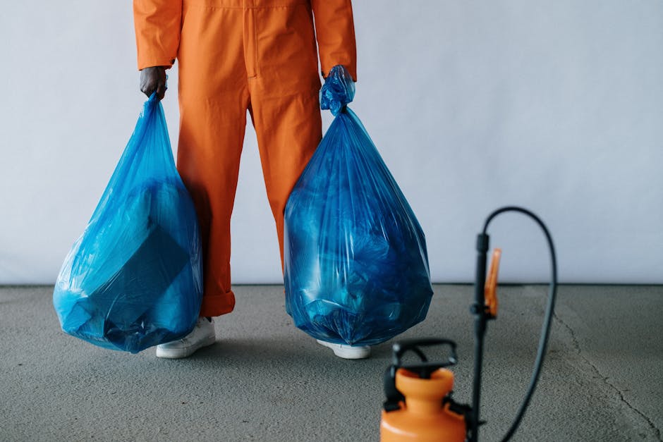 A person dressed in orange work overalls and white shoes is standing indoors on a gray concrete floor, holding two large blue plastic rubbish bags, each filled with waste and tied at the top. The individual's hands are gloved and gripping the bags, which are positioned in front of their legs. The background is a plain, light-colored wall, and in the foreground, there is a partially visible orange and black extension cord reel or power tool connected by a cable, indicating an environment related to waste collection or on-site clearance. The scene suggests an activity of rubbish collection, potentially part of a private waste removal service, with the bags and equipment prepared for disposal or transportation. This visual context aligns with professional rubbish removal practices, as offered by companies like Rubbish Removal Croydon, supporting the theme of bulky waste management and alternative disposal options in the Croydon area.